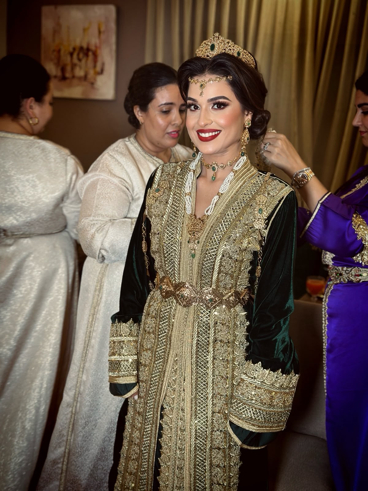 Mariée marocaine souriante en caftan brodé, entourée de préparatrices